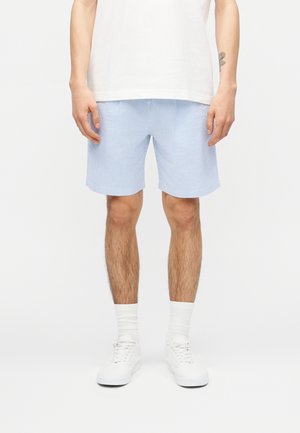 Homme portant un short à carreaux bleu clair, un t-shirt blanc, des chaussettes blanches et des baskets blanches, debout devant un fond uni.