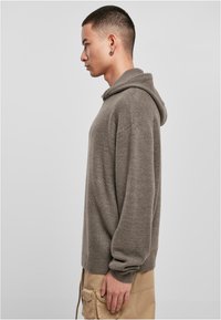 Khaki hooded sweater with a loose fit, ribbed cuffs, and a textured knit. Worn with beige cargo pants, shown in profile against a neutral background.