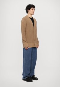 Beige oversized strikket cardigan med V-hals, frontknapper og lange ermer, kombinert med baggy blå denimjeans og svarte sko.