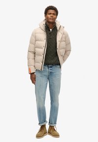 Beige pufferjakke med vandret quiltet design, grå kabelstrik sweater, lyseblå jeans og brune snørebåndsstøvler. Har orange accent.