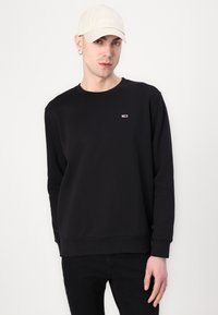 Sudadera negra de manga larga, cuello redondo y puños de canalé. Presenta un pequeño logo en rojo y blanco en el pecho. Lleva puesta una gorra clara.