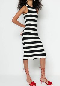 Nainen, joka on pukeutunut mustavalkoisiin vaakasuoriin raidallisiin hihattomiin midi-mekkoihin sekä punaisiin nauhakiilakorkoihin, jotka on koristeltu kukilla.