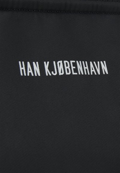 Fekete szövet sima textúrával, középen elhelyezve a "HAN KJØBENHAVN" felirat fehér, egyszerű, sans-serif betűtípussal.