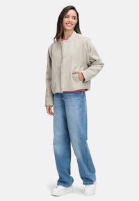 Hellgraue Bomberjacke aus strukturiertem Stoff mit Reißverschluss und seitlichen Taschen, kombiniert mit lockeren blauen Jeans und weißen Sneakers.