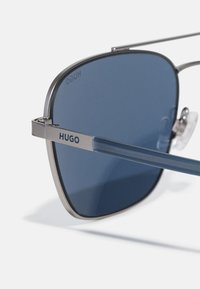 Ochelari de soare din metal cu un design unghiular, lentile de culoare albastru închis și ramă argintie, având brandingul "HUGO" pe bratul ochelarilor.