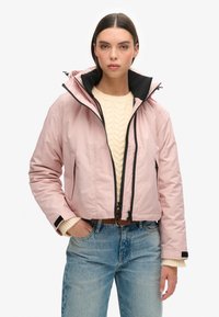Różowa kurtka typu cropped z czarnym wnętrzem, dwa przednie zamki błyskawiczne oraz elastyczne mankiety, noszona na kremowym swetrze i niebieskich dżinsach.
