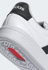 Zapatilla blanca de Adidas con lengüeta trasera negra, logo en relieve en la entresuela y acento rojo en la suela texturizada.