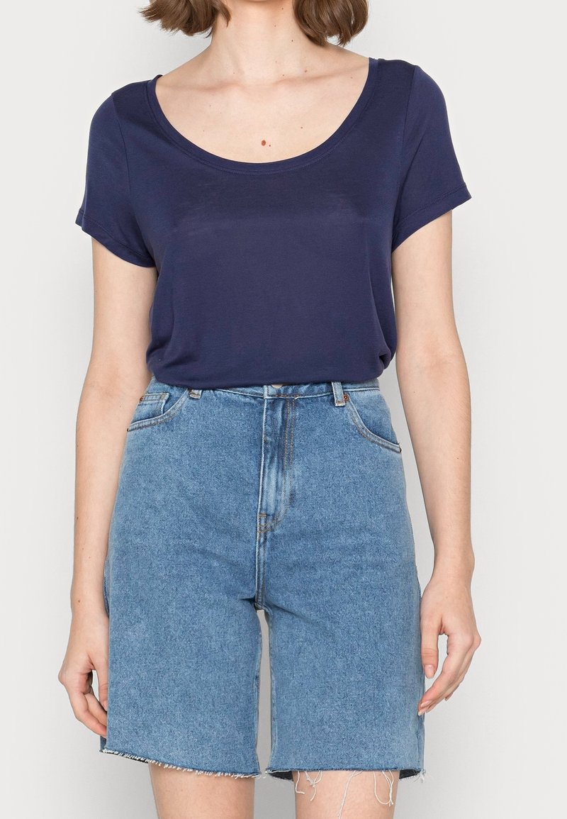 Femme portant un t-shirt bleu marine à col rond et manches courtes, rentré dans un short en jean bleu clair taille haute avec des bords effilochés.