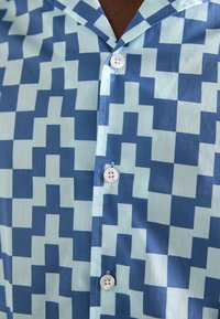 Κοντομάνικο πουκάμισο με κουμπιά σε γεωμετρικό μπλε και ανοιχτό μπλε σχέδιο, με γιακά και λευκά κουμπιά μπροστά.