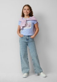 Lichtblauw T-shirt met een grafisch ontwerp, gecombineerd met lichtgewassen wijd-gesneden jeans. Een lila trui nonchalant over de schouders gedrapeerd.