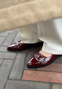 Glanzende bordeauxrode puntige schoenen hebben zilveren gesp-accenten en oogletdetails, gecombineerd met crèmekleurige wijde broekspijpen.