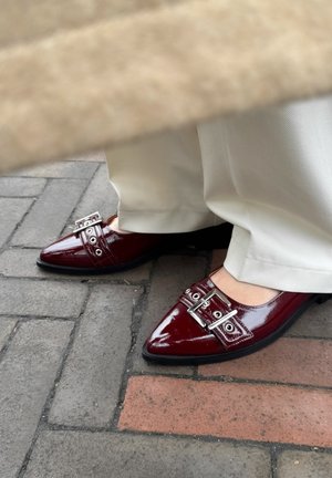 Glanzende bordeauxrode puntige schoenen hebben zilveren gesp-accenten en oogletdetails, gecombineerd met crèmekleurige wijde broekspijpen.