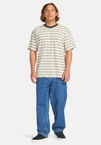 Striped t-shirt in cream with teal and navy horizontal lines, short sleeves, ribbed navy collar, and a small logo on the chest, paired with blue jeans.