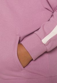 Sudadera con capucha de color púrpura claro con bolsillo tipo canguro. Presenta puños acanalados y una tela texturizada. Acento blanco a lo largo de la manga.