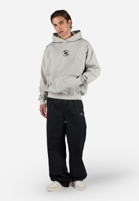 Lysegrå hættetrøje med en frontlomme og rund halsåbning, parret med sorte vide bukser og hvide sneakers. Logo printet på hættetrøjen.