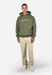 Felpa verde oliva con tasca anteriore e design stampato. Abbinata a pantaloni color crema e sneaker bianche. Vestibilità rilassata.