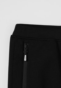 Un pantalon de survêtement en coton noir doté d'une taille élastique et d'une poche latérale zippée avec un tirette métallique. Tissu lisse avec une finition mate.