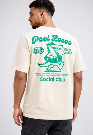 Mann trägt cremefarbenes T-Shirt mit türkisfarbiger Grafik eines laufenden Cartoon-Krokodils im Schwimmring und Schriftzug „Pool Locos Social Club, jeden Sonntag.“