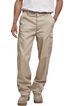Mann trägt beige Cargohose mit Seitentaschen, kombiniert mit braunen Arbeitsstiefeln und einem weißen Hemd, steht mit einer Hand in der Tasche.