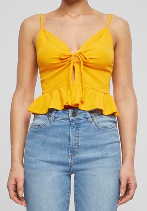 Crop top jaune sans manches avec lien devant et ourlet à volants, porté avec un jean taille haute bleu clair sur une personne devant un fond gris.