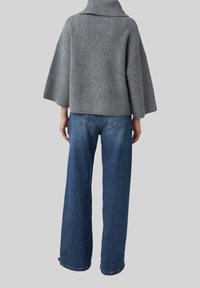 Personne portant un pull à col roulé côtelé gris aux manches larges et un jean large bleu, se tenant devant un fond uni.