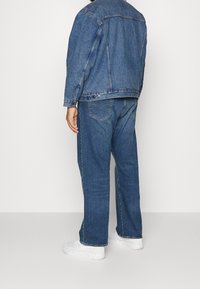 Man som bär en blå denimjacka och jeans med vita sneakers, står vänd bortåt mot en enfärgad bakgrund.