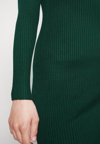 Mörkgrön ribbad stickad tyg som visar upp förlängda ärmar och en figurnära silhuett. Texturen är slät med subtila glansiga detaljer.