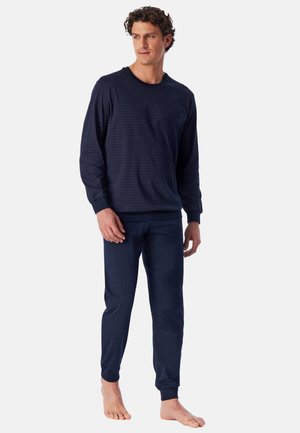 Man staat op blote voeten, draagt een donkerblauw shirt met lange mouwen en bijpassende broek tegen een effen lichte achtergrond.