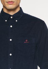Camicia in corduroy blu navy con colletto a bottoni, dotata di taschino sul petto a sinistra e logo ricamato rosso. Tessuto testurizzato con righe verticali.