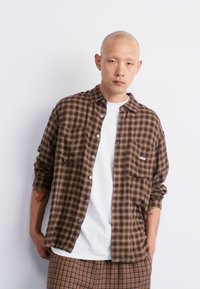Brown checkered button-up shirt with a chest pocket, layered over a white t-shirt. Model wears patterned pants, with short sleeves and a collar.