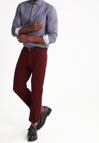 Camicia grigia con maniche lunghe, abbinata a pantaloni bordeaux e scarpe nere. La camicia ha una tasca e le maniche sono arrotolate.