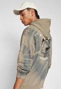 Haut à capuche tie-dye vert olive avec un design graphique au dos, doté d'une poche kangourou et d'une capuche réglable, associé à une casquette blanche.