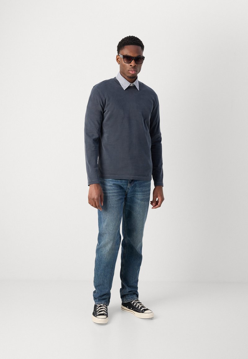 Dunkelblaues Langarmshirt über einem gestreiften Hemd mit Kragen, kombiniert mit hellblauen Jeans und schwarzen Turnschuhen, getragen von einem Mann mit Sonnenbrille.