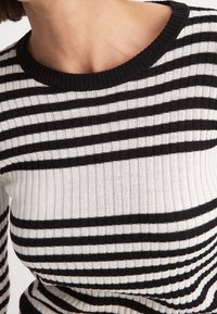 Gros plan sur une personne portant un pull en maille côtelée à rayures horizontales noires et blanches avec un col rond.