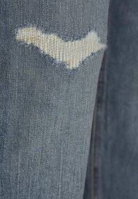 Jeans de mezclilla azul con una sección desgastada y bordes deshilachados, además de un parche de color más claro que revela la textura de la tela subyacente.
