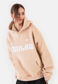 Beżowa, oversize'owa bluza z kapturem wykonana z teksturowanej tkaniny. Posiada przednią kieszeń oraz białe, wyszywane litery z dekoracyjnymi liniami.