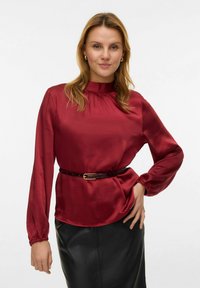 Bordeauxrode satijnen blouse met lange mouwen, hoge halslijn en geplooide details, in de taille aangesnoerd met een dunne, donkere riem.