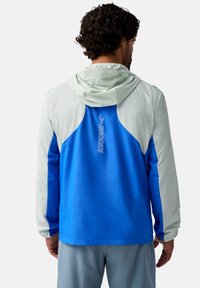 Kapuzensportjacke mit blauem Körper und hellgrauen Akzenten. Das Material wirkt leicht, hat eine glatte Textur und ein Logo-Detail.