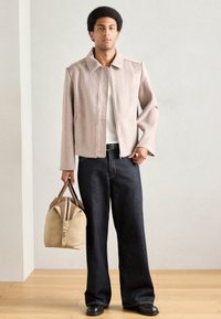 Beżowa kurtka na zamek z kołnierzem, zestawiona z ciemnoniebieskimi jeansami o szerokich nogawkach. Do zestawu dołączona jest beżowa torba typu duffel z ciemnymi akcentami.