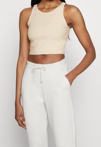 Femme noire portant un crop top beige sans manches et un pantalon de jogging blanc avec cordon, une main dans la poche, sur un fond uni.