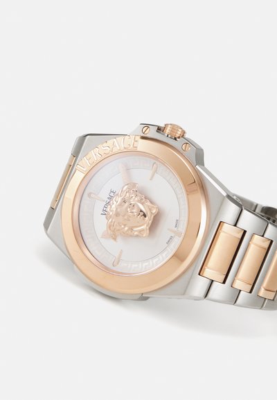 Versace laikrodis su aštuonkampiu sidabro ir rožinio aukso metaliniu korpusu, turintis baltą ciferblatą, liūto emblema ir sujungtą apyrankę.