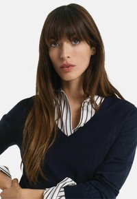 Marineblauwe V-hals trui laagjes over een witte blouse met dunne marineblauwe strepen. Heeft lange mouwen en een lichte textuur in de brei.