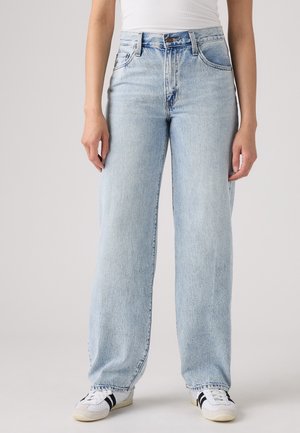 Person trägt hellblaue, hoch taillierte Jeans mit weitem Bein, kombiniert mit weißen Sneakers mit schwarzen Streifen und einem weißen Oberteil.