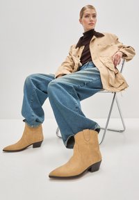 Botine scurte din suede bej, cu vârful ascuțit, detalii subtile de cusătură și toc mic, asortate cu blugi evazați și o jachetă bej.