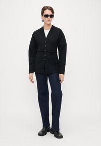 Svart, knappet cardigan med belte i midjen, med V-hals og lange ermer. Kombinert med mørkeblå jeans og kraftige svarte støvler.