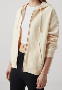 Mujer vistiendo una sudadera ligera beige con cremallera sobre una camiseta blanca sin mangas y pantalones negros con cordón, de pie con un brazo relajado.