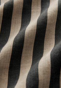 Pellava kangas, jossa vuorottelevat mustat ja beige raidat, esittelee teksturoidun pinnan, jossa näkyy kuituja ja aaltoilevaa roikkumista.