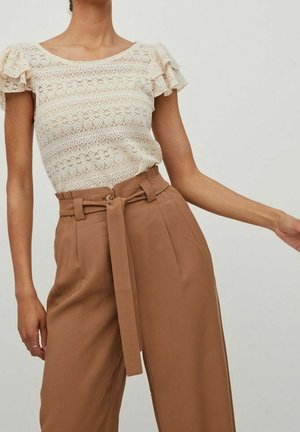 Kobieta w kremowej koronkowej bluzce z krótkim rękawem, włożonej do wysokich brązowych spodni o szerokich nogawkach, przewiązanych pasem z tego samego materiału.