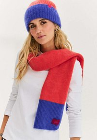 Czerwono-niebieski dziergany szalik o prążkowanej fakturze; jednolite bloki kolorów; noszony z prostą białą bluzką i dopasowaną czapką beanie.
