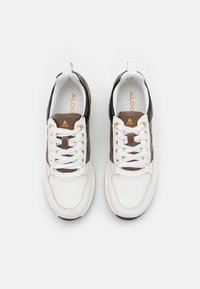 Vita och bruna sneakers med en texturerad ovandel, svarta accenter och vita snören. Guldlogotyp på tungan och kontrasterande sula.